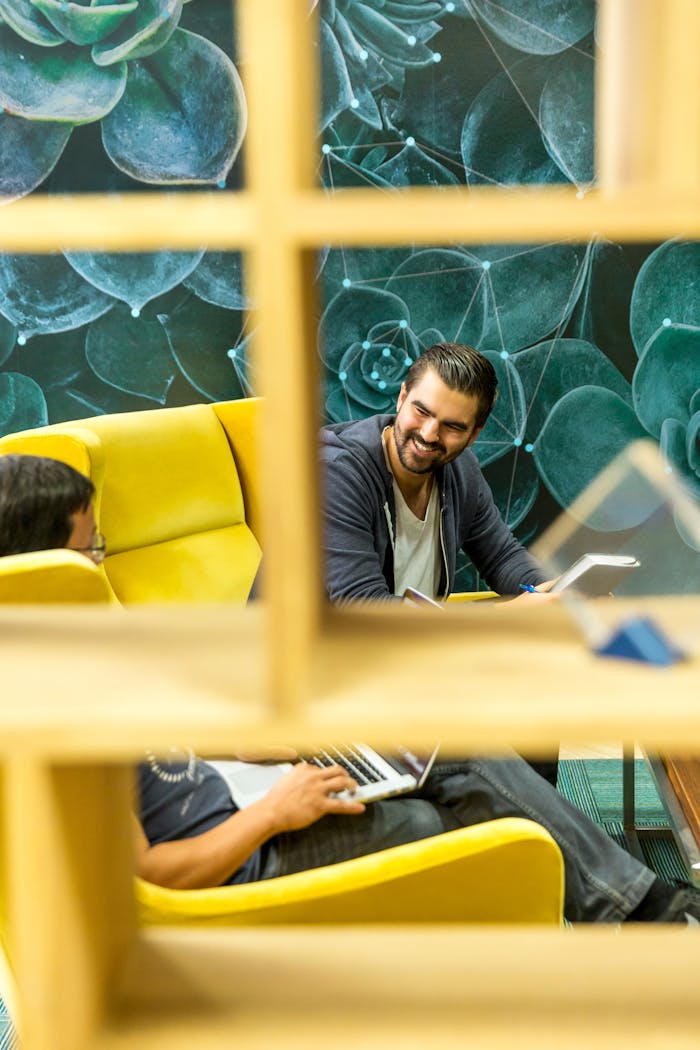 Window With the View of Man Holding Book While Sitting Beside Man Using Laptop